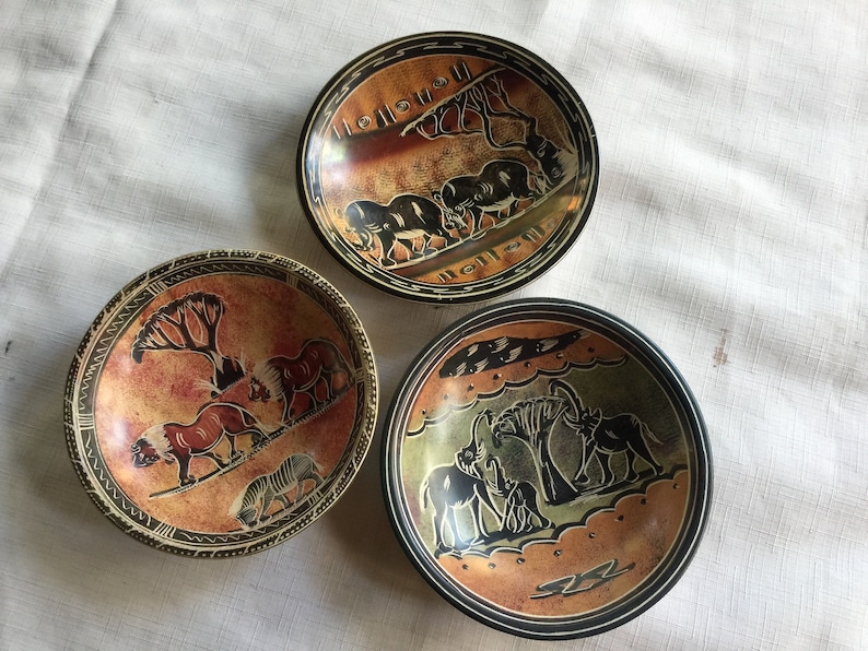 May include: Three small, round, decorative bowls with black and brown designs of African animals and trees. The bowls are made of a dark, textured material, possibly slate.