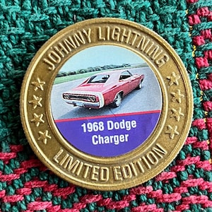 Puede incluir: Una moneda dorada de edición limitada de Johnny Lightning con un Dodge Charger rojo de 1968. La moneda tiene un centro circular con la imagen del coche y texto. El borde de la moneda tiene las palabras "JOHNNY LIGHTNING" y estrellas.