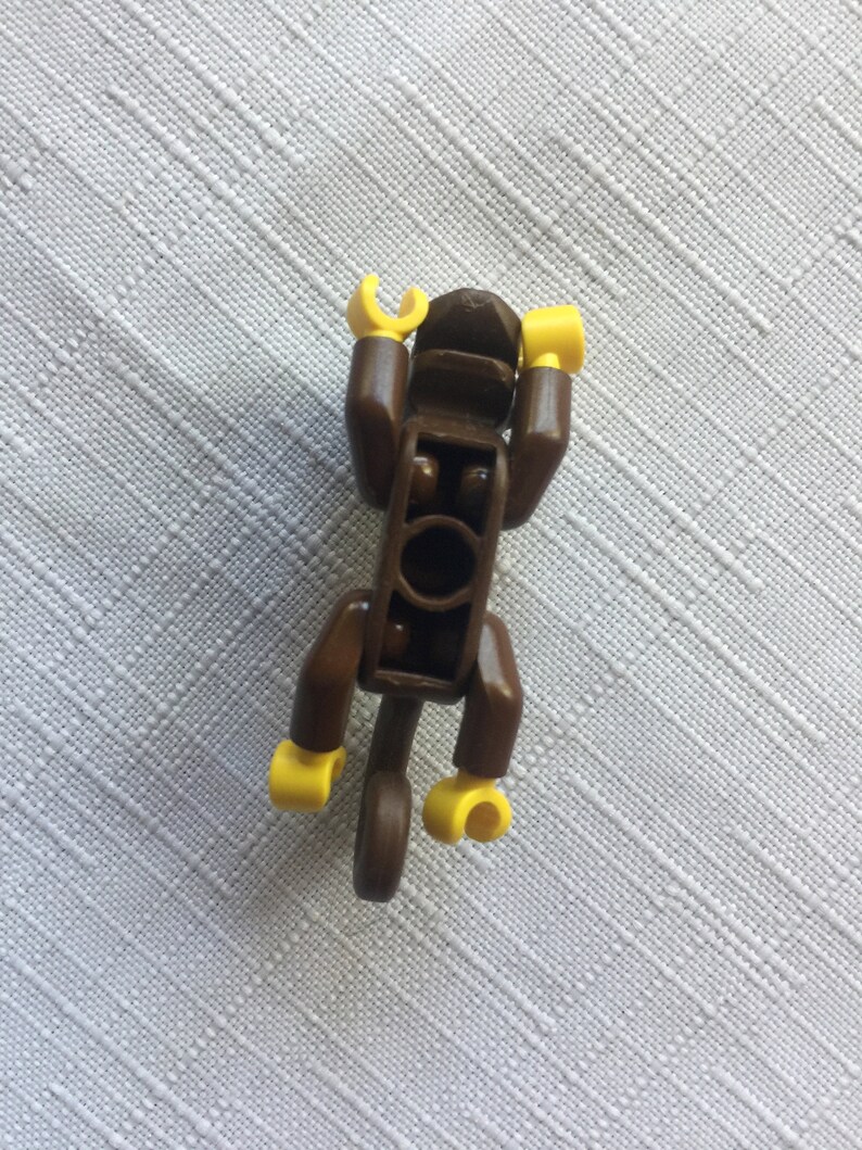 May include: A brown Lego monkey figure with yellow hands and feet. The monkey is in a sitting position with its arms raised above its head.