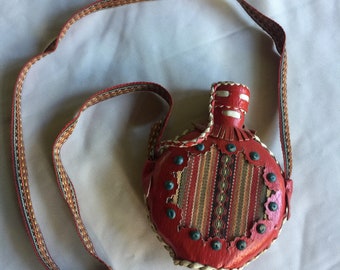 Peut inclure: Une gourde en cuir rouge avec un motif tissé et une sangle tressée. La gourde a un détail de frange et est décorée de perles bleues et blanches.