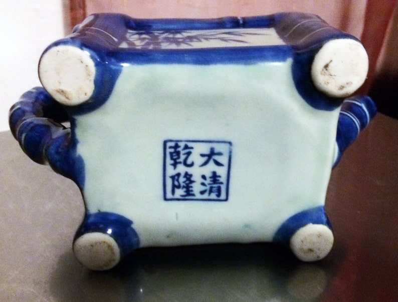 Peut inclure: Un pot en c&eacute;ramique bleu et blanc avec quatre pieds et une base carr&eacute;e. Le pot est d&eacute;cor&eacute; de branches et de feuilles de bambou. Le fond du pot porte un tampon carr&eacute; avec le texte "敬大 曜清" en caract&egrave;res chinois.