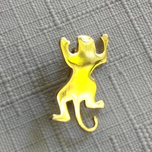 May include: A gold-colored metal monkey pin with a raised design. The monkey is in a climbing pose with its arms raised above its head.