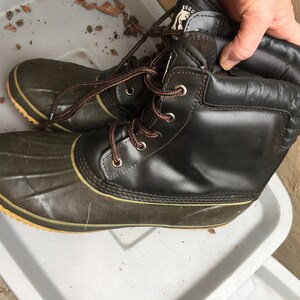 May include: A pair of brown leather winter boots with green rubber bottoms and brown laces. The boots have a yellow trim around the bottom and a brown leather cuff.