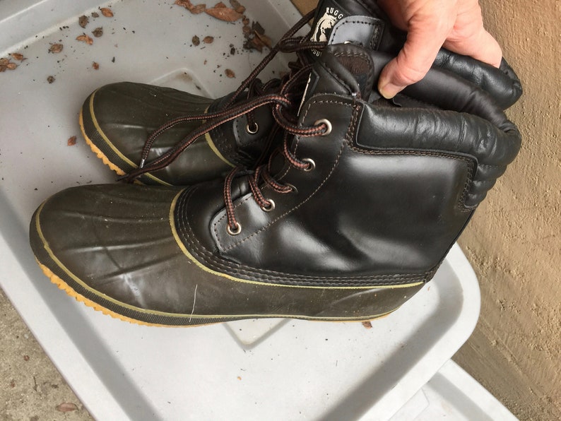 May include: A pair of brown leather winter boots with green rubber bottoms and brown laces. The boots have a yellow trim around the bottom and a brown leather cuff.