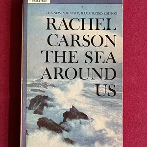 Puede incluir: Una portada de libro de bolsillo vintage en azul y blanco. El título es "Rachel Carson El Mar Que Nos Rodea" con las palabras "The Newly Revised, Illustrated Edition" sobre el título. La portada presenta una fotografía de una costa rocosa con olas rompiendo.