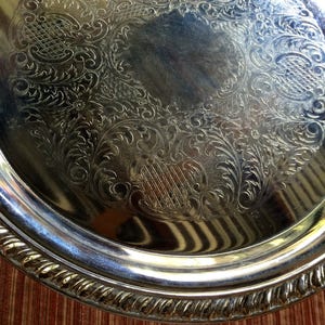 May include: A silver serving tray with an ornate, embossed design. The tray has a raised rim and a smooth, reflective surface.