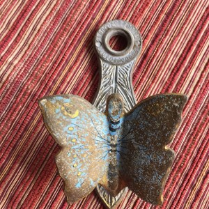 May include: A vintage metal butterfly-shaped wall hook. The butterfly has blue and gold wings with a detailed design. The hook is attached to a decorative metal piece with a circular opening at the top.