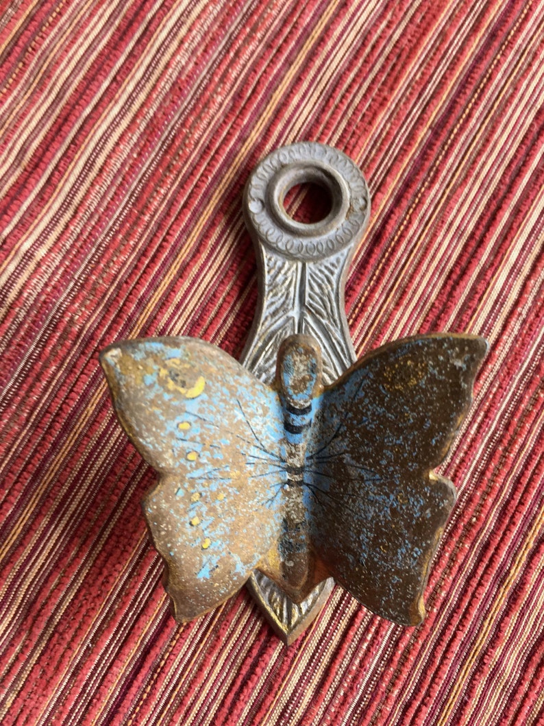 May include: A vintage metal butterfly-shaped wall hook. The butterfly has blue and gold wings with a detailed design. The hook is attached to a decorative metal piece with a circular opening at the top.