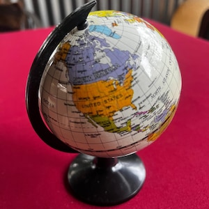 Op de afbeelding: Een kleine, kleurrijke wereldbol met een zwarte standaard en een zwarte meridiaanring. De wereldbol toont een gedetailleerde wereldkaart, met continenten in verschillende kleuren. De basis is rond en glanzend. De wereldbol staat tegen een rode achtergrond.
