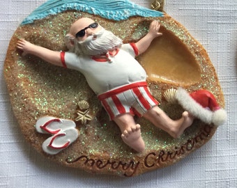 以下が含まれることがあります： ビーチでくつろぐサンタクロースを描いたクリスマスオーナメント。サンタはサングラス、赤いトリムの白いシャツ、赤と白のストライプのショーツを着用しています。オーナメントには小さなサンタ帽子、ビーチサンダル、そして「merry christmas」の文字が含まれています。