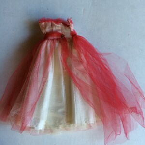 May include: A vintage doll dress featuring a cream-colored bodice and a red tulle overlay. The dress has a satin-like top with a small button detail and a flowing skirt. The tulle creates a layered effect, adding volume and texture to the garment.