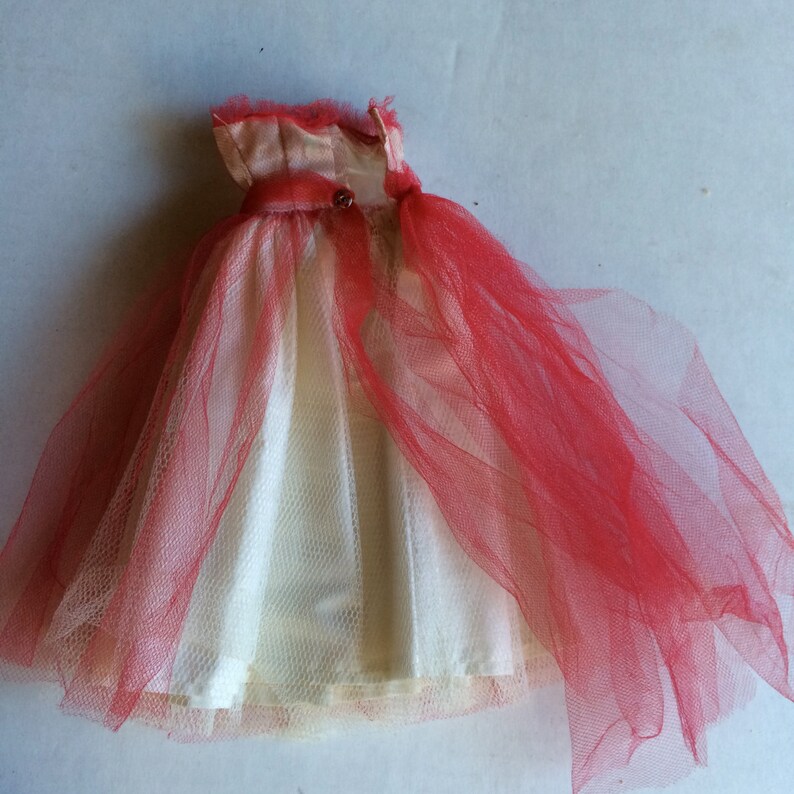 May include: A vintage doll dress featuring a cream-colored bodice and a red tulle overlay. The dress has a satin-like top with a small button detail and a flowing skirt. The tulle creates a layered effect, adding volume and texture to the garment.