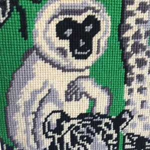 May include: A close-up of a needlepoint tapestry featuring a white monkey with a black face and a black and white tiger. The background is green.