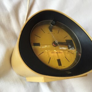 May include: A yellow and black vintage Panasonic alarm clock with a round face and a black bezel. The clock has a white background with black hour and minute hands and a red second hand. The clock is set to 10:51.