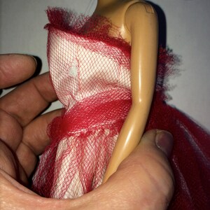 May include: A doll wearing a red and white dress. The dress features a red mesh overlay and a ruffled neckline. The doll is held by a person's hand, showcasing the dress's design and texture.