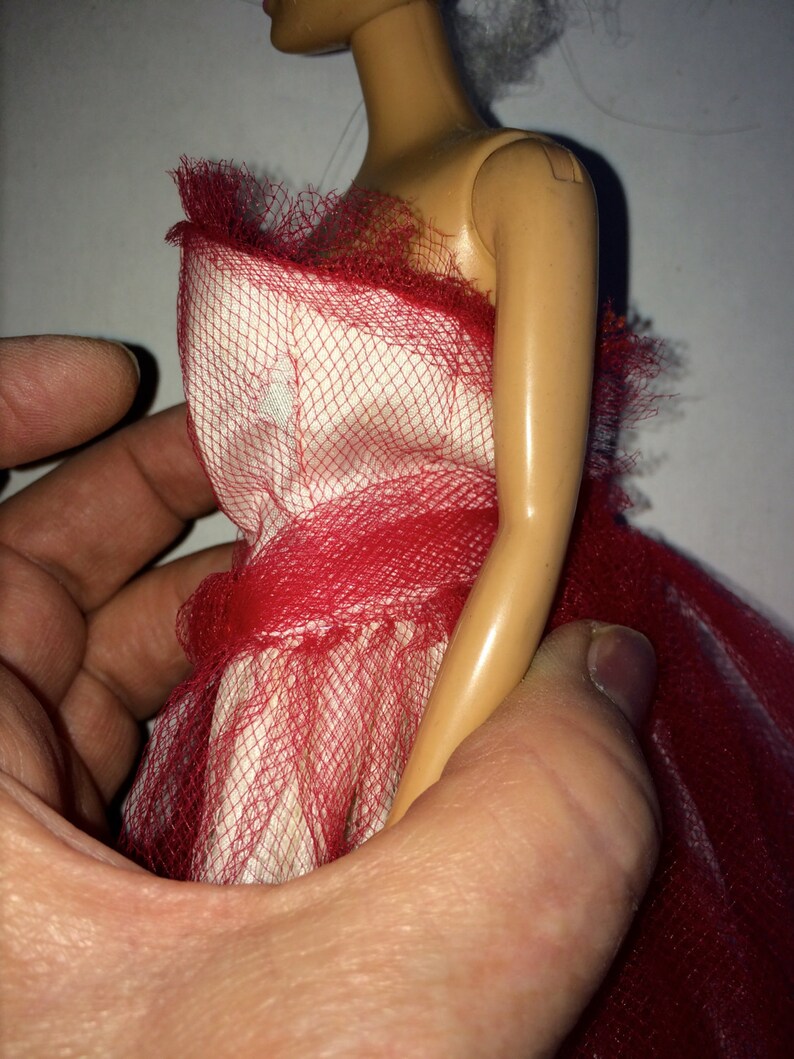 May include: A doll wearing a red and white dress. The dress features a red mesh overlay and a ruffled neckline. The doll is held by a person's hand, showcasing the dress's design and texture.