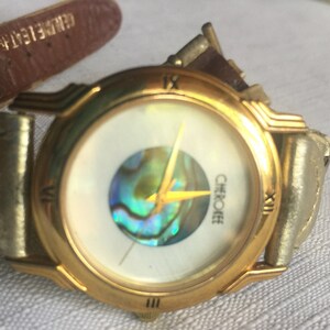 May include: A gold-tone wristwatch with a white face and a mother-of-pearl inlay. The watch has a brown leather band and the word "Cherokee" is printed on the face.