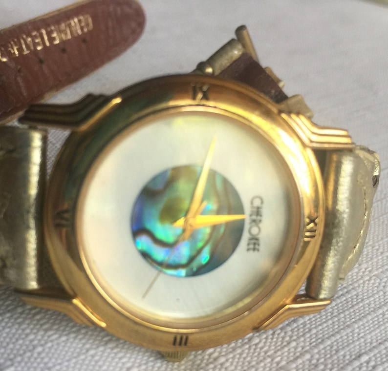 May include: A gold-tone wristwatch with a white face and a mother-of-pearl inlay. The watch has a brown leather band and the word "Cherokee" is printed on the face.