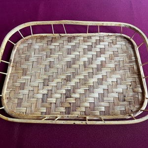 May include: Rectangular woven bamboo tray with a light brown and tan color scheme. The tray has a raised border with a handle-like design on the sides. The woven pattern is tightly knit, creating a textured surface.