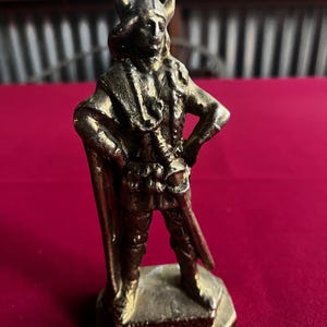 May include: A gold-colored metal figurine of a Viking warrior wearing a horned helmet and holding a sword. The figurine is standing on a square base.