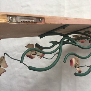 May include: A wooden shelf with a green metal floral design. The shelf is attached to the wall with a metal bracket. The floral design features pink and white flowers.
