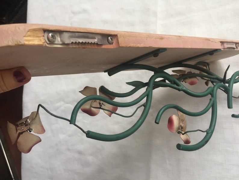 May include: A wooden shelf with a green metal floral design. The shelf is attached to the wall with a metal bracket. The floral design features pink and white flowers.