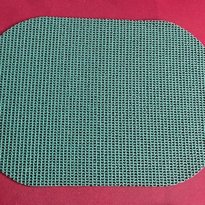 Op de afbeelding: Een ovaal turquoise matje met een geweven textuur, geplaatst tegen een effen rode achtergrond. Het ontwerp van de mat suggereert dat deze gebruikt kan worden als keuken- of badkameraccessoire.