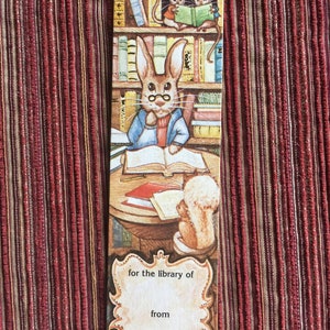 Peut inclure: Un marque-page représentant un lapin portant des lunettes et lisant un livre dans un décor de bibliothèque. Deux souris lisent également des livres sur une étagère. Le marque-page comporte un espace vide pour "pour la bibliothèque de" et "de la part de".