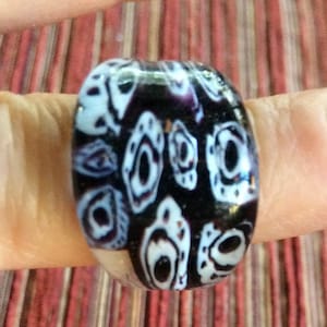 May include: A black and white glass ring with a swirling pattern. The ring is on a finger.