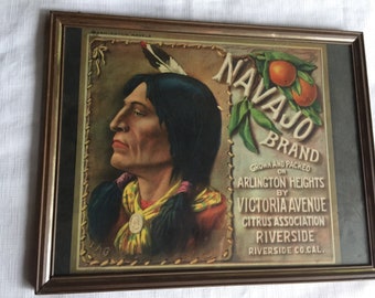 May include: A vintage framed advertisement for Navajo Brand oranges. The ad features a Native American man in profile, wearing a feathered headdress and a colorful scarf. The text reads "Navajo Brand", "Grown and Packed on Arlington Heights by Victoria Avenue Citrus Association Riverside Riverside Co.Cal."
