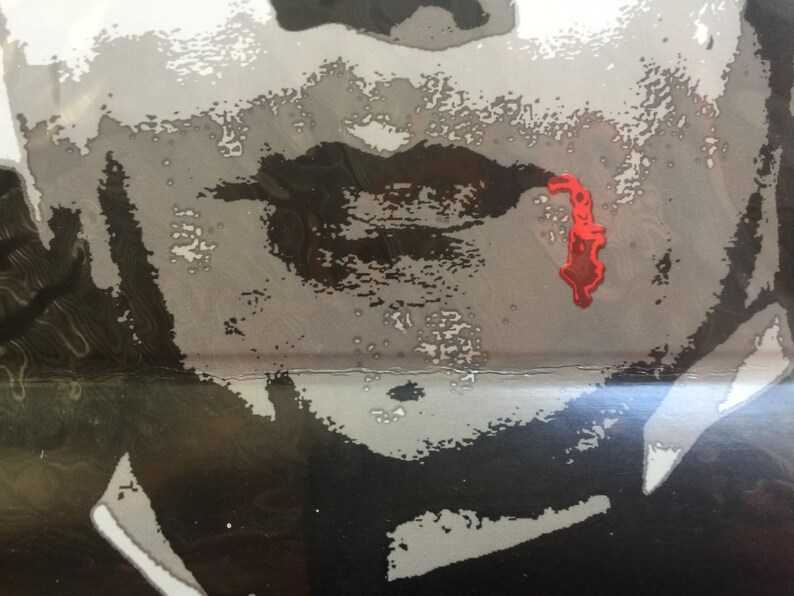 May include: A close-up of a person's face with a black and white filter. The person's lips are painted black and there is a red splatter of paint on their cheek.