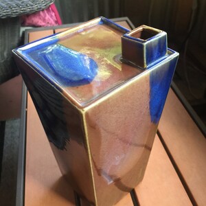 Op de afbeelding: Een vierkante keramische vaas met een unieke tuit. De vaas is geglazuurd in tinten bruin, blauw en goud, wat een veelkleurig effect cre&euml;ert.