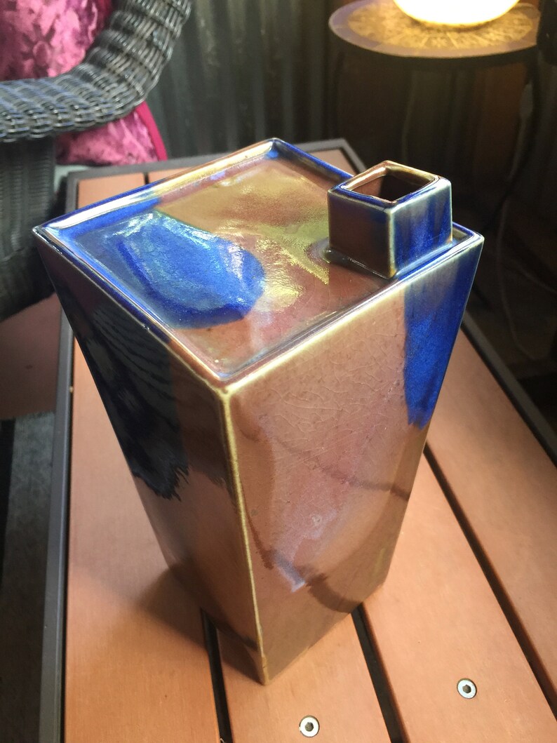 Op de afbeelding: Een vierkante keramische vaas met een unieke tuit. De vaas is geglazuurd in tinten bruin, blauw en goud, wat een veelkleurig effect cre&euml;ert.