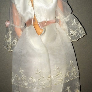 Puede incluir: Un vestido blanco de mu&ntilde;eca con una cinta de raso rosa alrededor de la cintura. El vestido tiene una superposici&oacute;n transparente con un delicado borde de encaje y bordados florales.