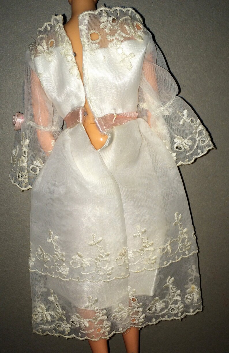 Puede incluir: Un vestido blanco de mu&ntilde;eca con una cinta de raso rosa alrededor de la cintura. El vestido tiene una superposici&oacute;n transparente con un delicado borde de encaje y bordados florales.