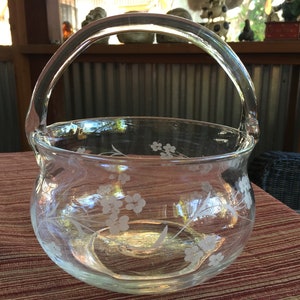 May include: A clear glass bowl with a handle, etched with a floral design. The bowl is sitting on a red and brown patterned surface.