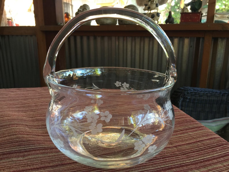 May include: A clear glass bowl with a handle, etched with a floral design. The bowl is sitting on a red and brown patterned surface.
