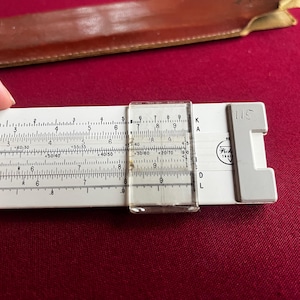 May include: A white slide rule with black markings and a clear plastic cursor. The slide rule has a white plastic frame and a gray end piece with the number "115". A brown leather case is in the background.