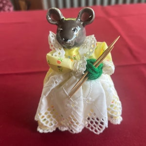 May include: A ceramic mouse figurine wearing a yellow dress with white lace trim and holding knitting needles and yarn.