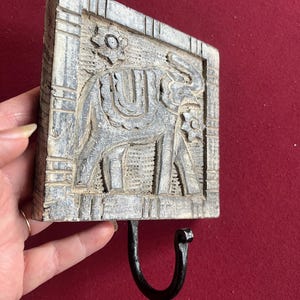 May include: A gray, rectangular wooden wall hook with a carved elephant design. The elephant is centered within a rectangular frame, and a black metal hook is attached below. Floral accents are carved at the top and bottom of the frame.