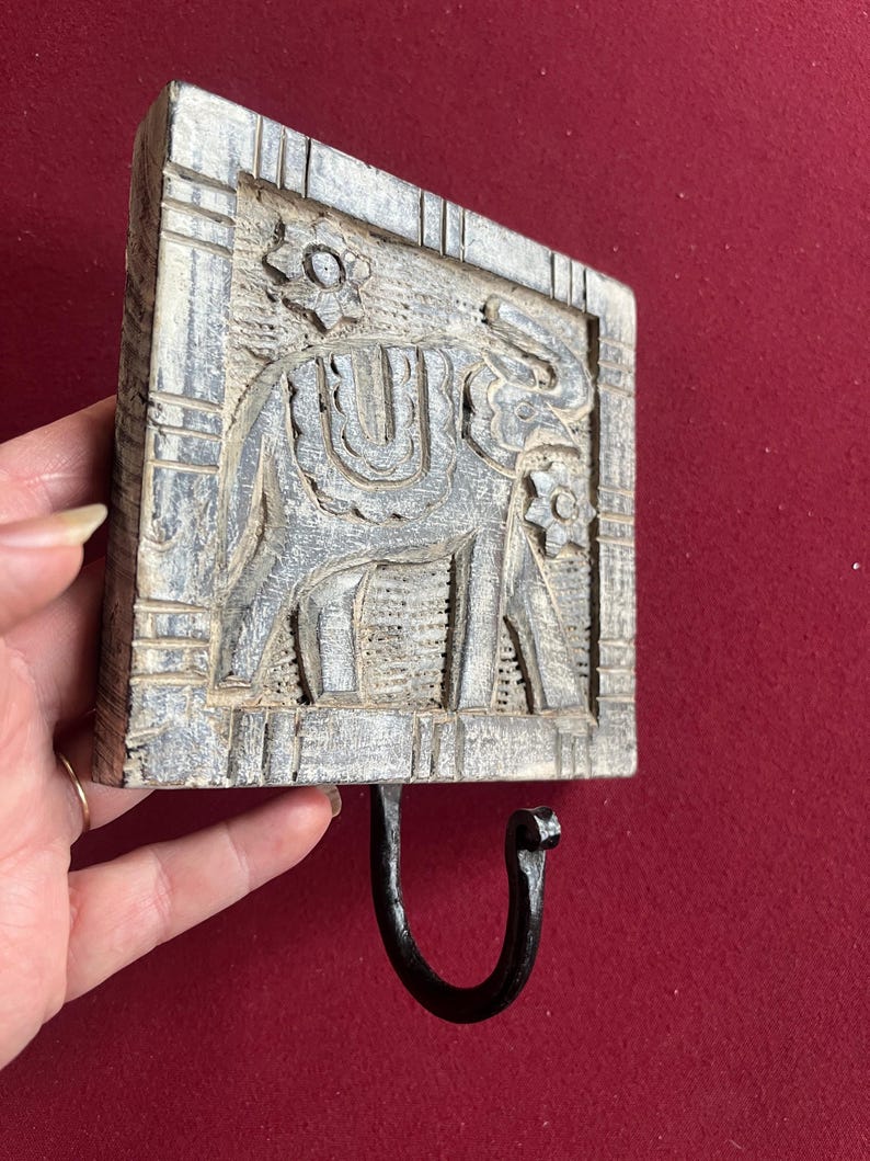 May include: A gray, rectangular wooden wall hook with a carved elephant design. The elephant is centered within a rectangular frame, and a black metal hook is attached below. Floral accents are carved at the top and bottom of the frame.