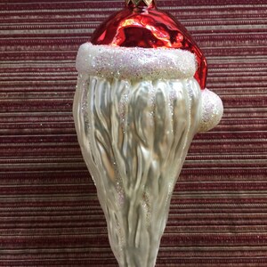 以下が含まれることがあります： サンタクロースの形をしたクリスマスオーナメント。光沢のある赤い帽子、白いキラキラのひげ、吊り下げ用の金色の金属ループが特徴です。背景は、バーガンディと白のストライプの生地です。