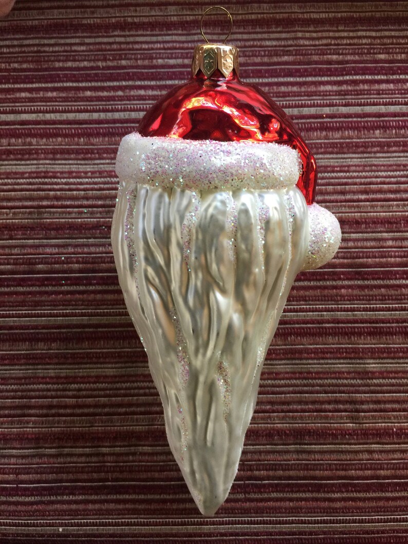 以下が含まれることがあります： サンタクロースの形をしたクリスマスオーナメント。光沢のある赤い帽子、白いキラキラのひげ、吊り下げ用の金色の金属ループが特徴です。背景は、バーガンディと白のストライプの生地です。