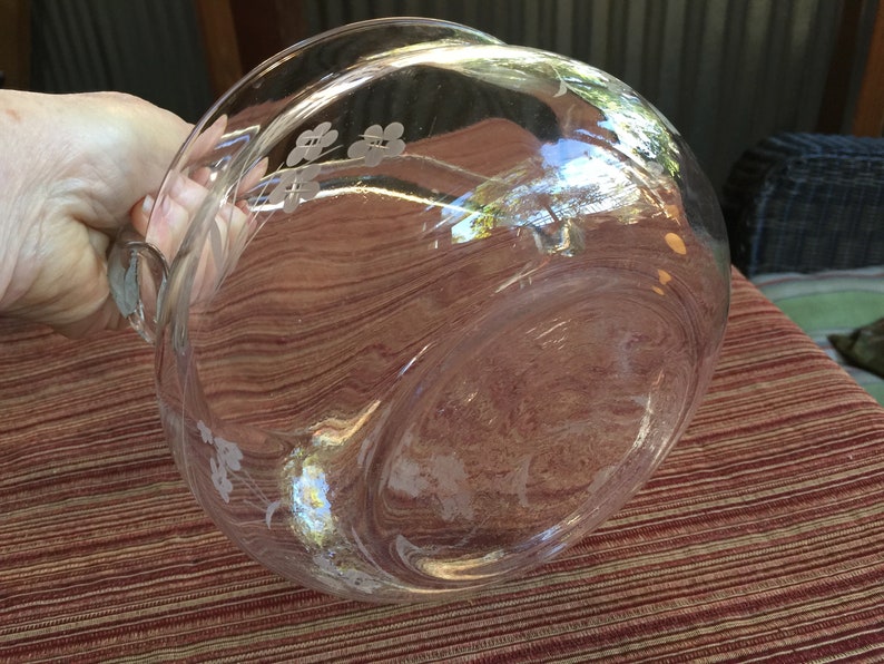 May include: A clear glass serving bowl with a floral design etched into the side. The bowl has a handle and is sitting on a red and brown patterned surface.