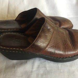 May include: Brown leather slip-on clogs with a wedge heel. The clogs have a stitched seam across the top and the brand name "Born" is visible on the insole.