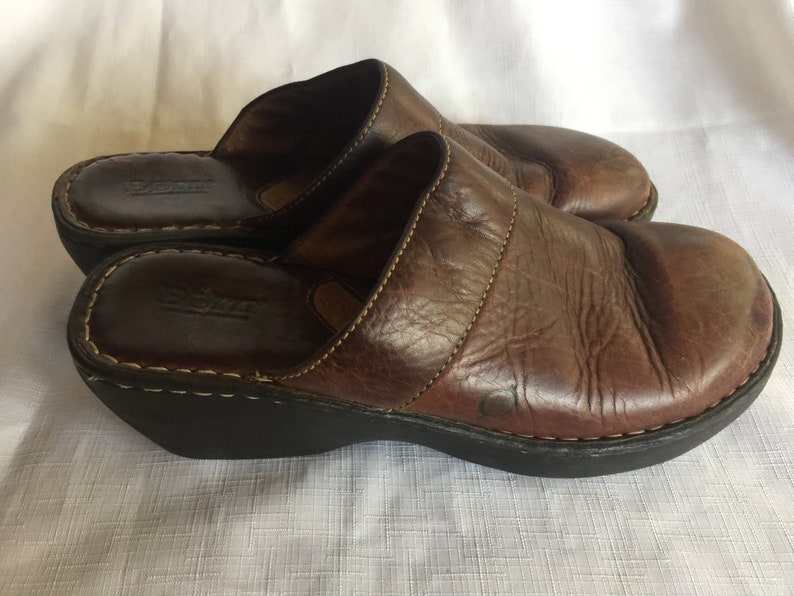 May include: Brown leather slip-on clogs with a wedge heel. The clogs have a stitched seam across the top and the brand name "Born" is visible on the insole.
