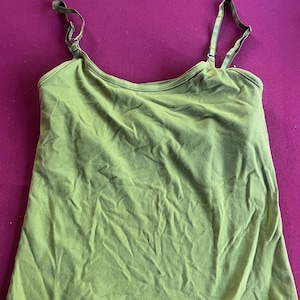 May include: Olive green camisole top with adjustable straps. The top is made of a soft, slightly wrinkled fabric. The straps are a darker shade of green and have a camouflage pattern.