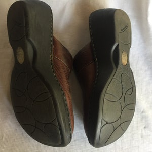 May include: A pair of brown leather clogs with a black rubber sole. The soles have a circular pattern.