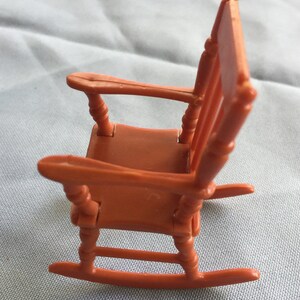 K&ouml;nnte beinhalten: Ein Miniatur-Schaukelstuhl aus Kunststoff, in einem warmen Orangeton lackiert. Der Stuhl hat ein schlichtes Design mit einer geschwungenen R&uuml;ckenlehne und einer breiten Sitzfl&auml;che. Die Schaukelbasis ist sichtbar und tr&auml;gt zum Charme dieses Miniaturst&uuml;cks bei.