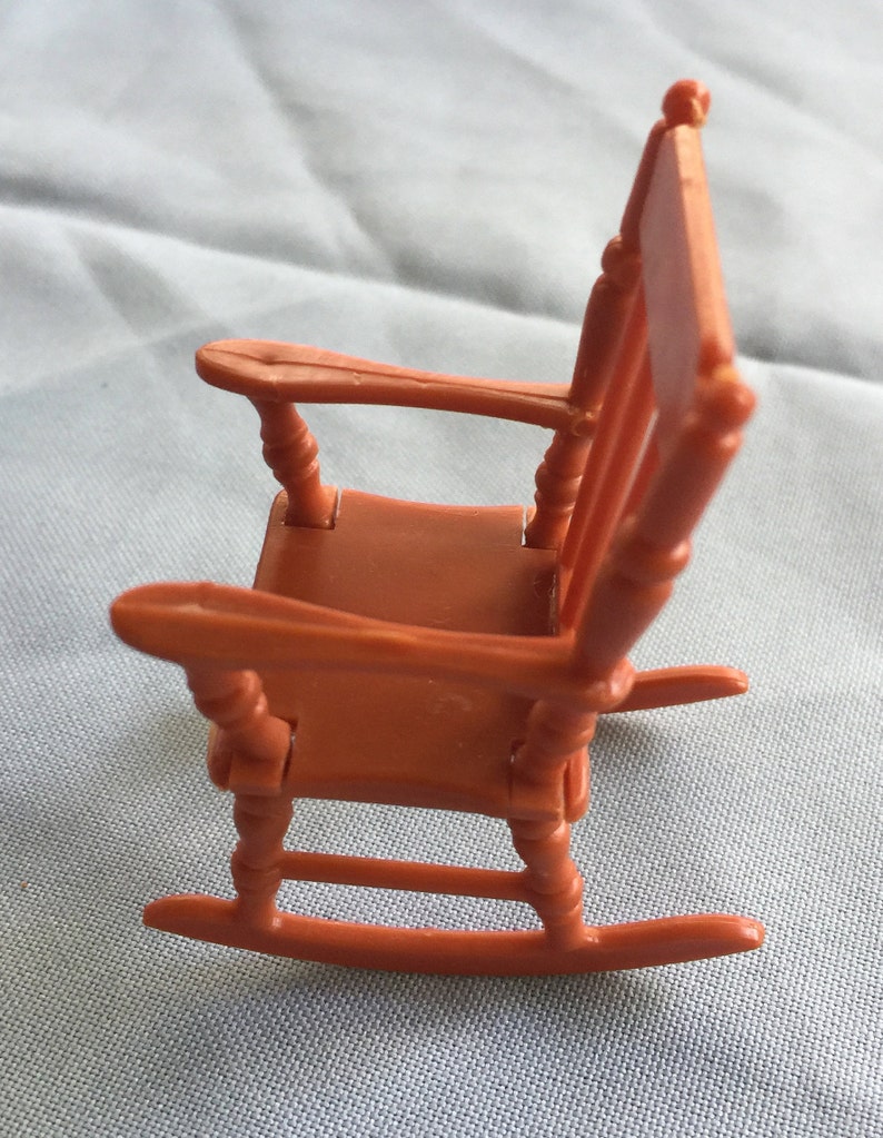 K&ouml;nnte beinhalten: Ein Miniatur-Schaukelstuhl aus Kunststoff, in einem warmen Orangeton lackiert. Der Stuhl hat ein schlichtes Design mit einer geschwungenen R&uuml;ckenlehne und einer breiten Sitzfl&auml;che. Die Schaukelbasis ist sichtbar und tr&auml;gt zum Charme dieses Miniaturst&uuml;cks bei.